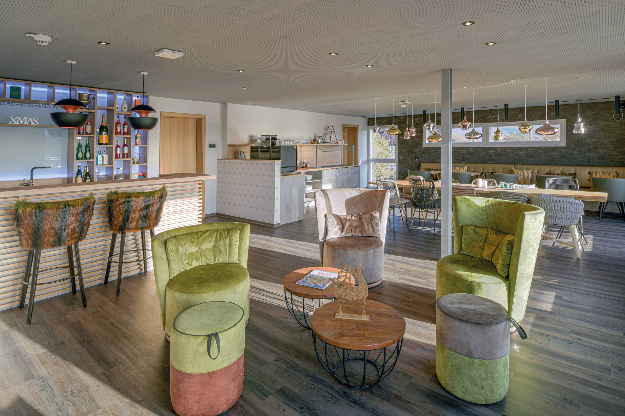 Eine moderne, helle Lounge mit einer Bar im Hintergrund. Es gibt gemütliche Sitzgelegenheiten, darunter zwei grüne Sessel, eine grau-beige Couch und runde Holztische. Hinter der Bar sind Regale mit verschiedenen Getränken. Das Ambiente ist einladend und stylisch.