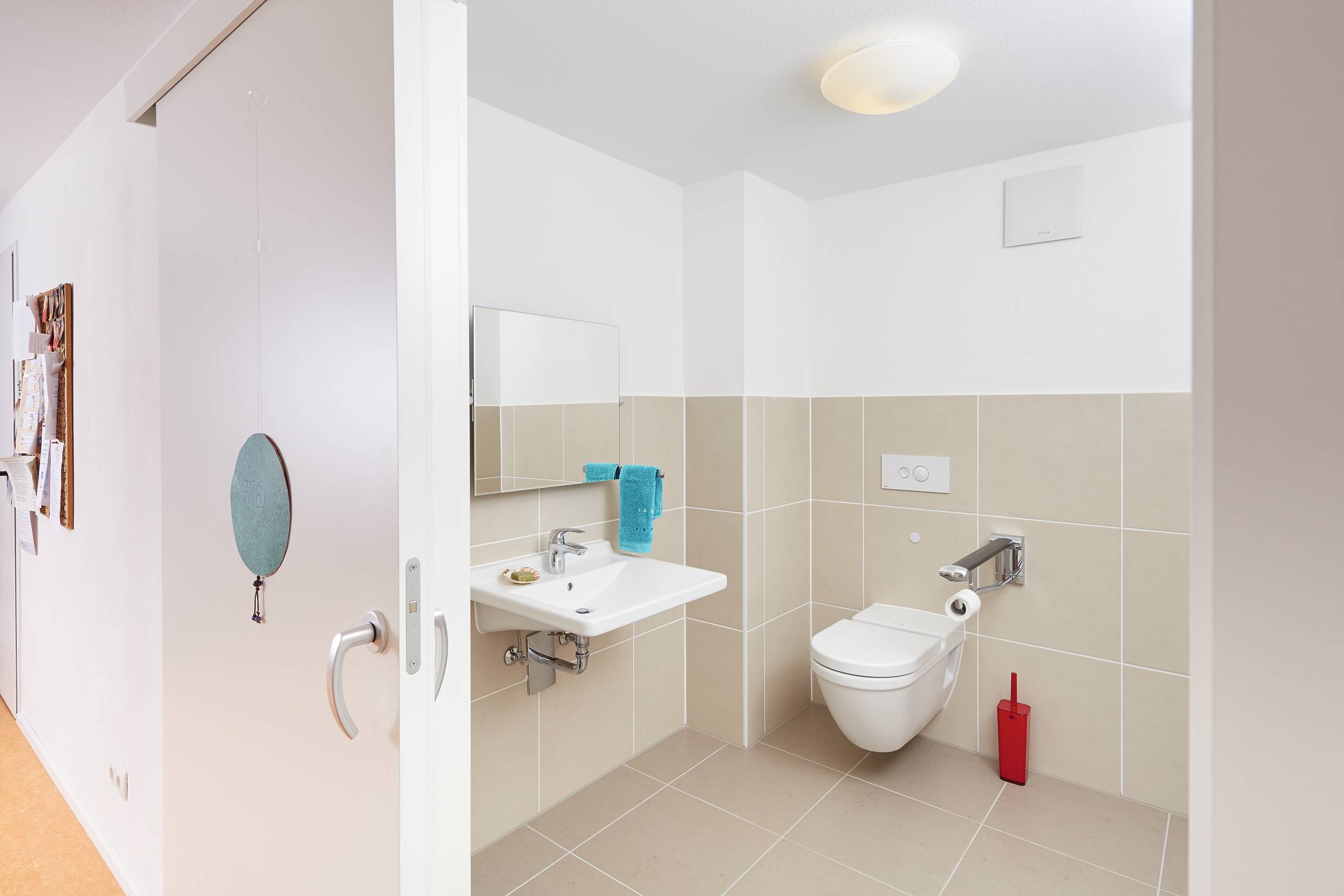 Das Bild zeigt ein barrierefreies Badezimmer mit einem Waschbecken, einem Spiegel und einer Toilette. An der Wand hängt ein türkisfarbener Handtuch. Der Boden ist hell und die Wände sind in neutralen Farbtönen gefliest. Die Tür ist teilweise offen.