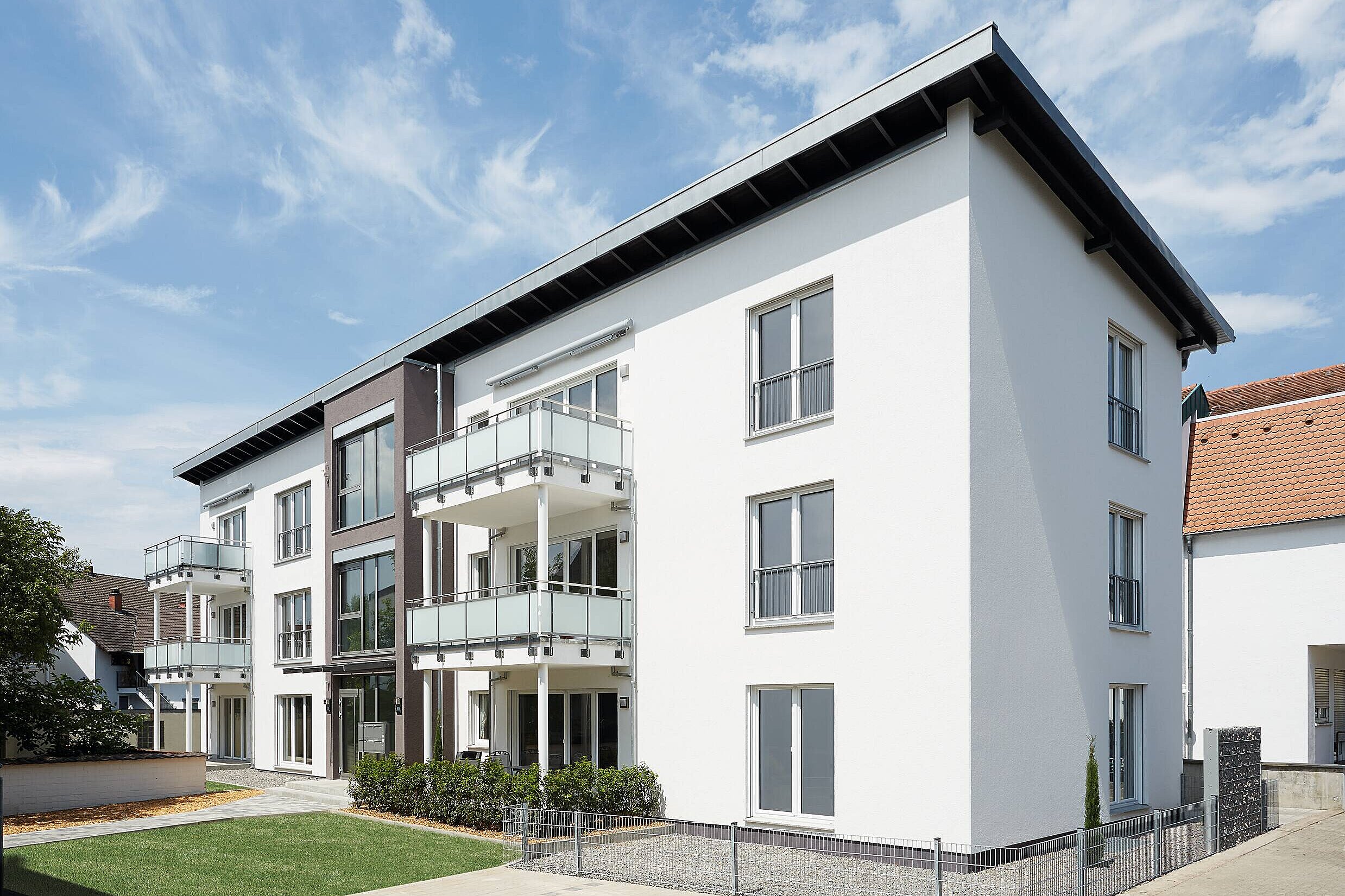 Ein modernes Mehrfamilienhaus mit weißen Wänden und großen Fenstern. Balkone aus Glas befinden sich an der Vorderseite. Im Vordergrund liegt eine gepflegte Grünfläche. Der Himmel ist blau mit einigen Wolken. Das Gebäude strahlt Helligkeit und Offenheit aus.