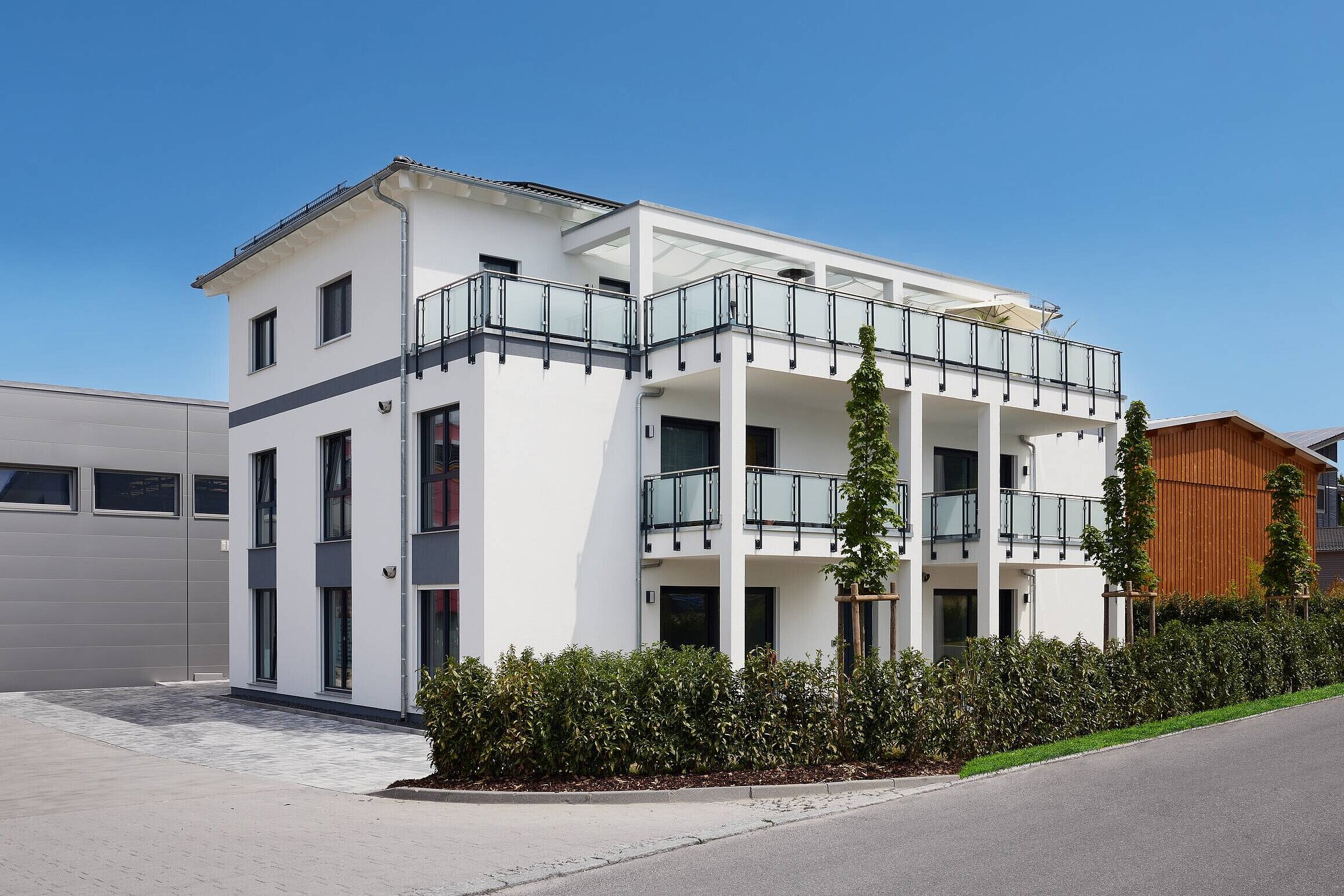 Ein modernes, dreigeschossiges Wohnhaus in Weiß mit Balkonen aus Glas. Die Fassade hat große Fenster und graue Akzente. Im Vordergrund eine gepflasterte Zufahrt und grünes Gestrüpp. Im Hintergrund ist ein graues und ein holzverkleidetes Gebäude sichtbar. Der Himmel ist blau und klar.