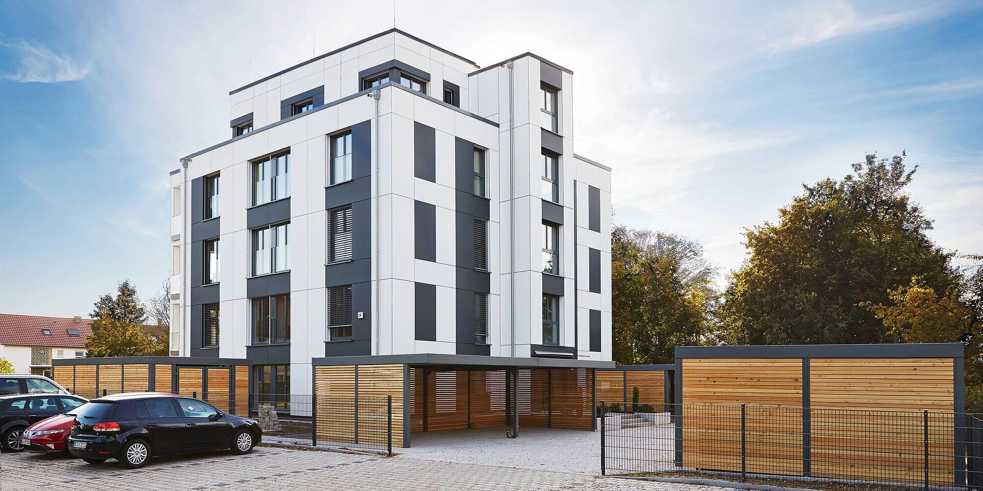 Ein modernes Mehrfamilienhaus mit weißen und grauen Fassadenelementen. Vor dem Gebäude befinden sich Parkplätze für Autos. Im Hintergrund sind Bäume erkennbar. Der Himmel ist klar und leicht bewölkt.