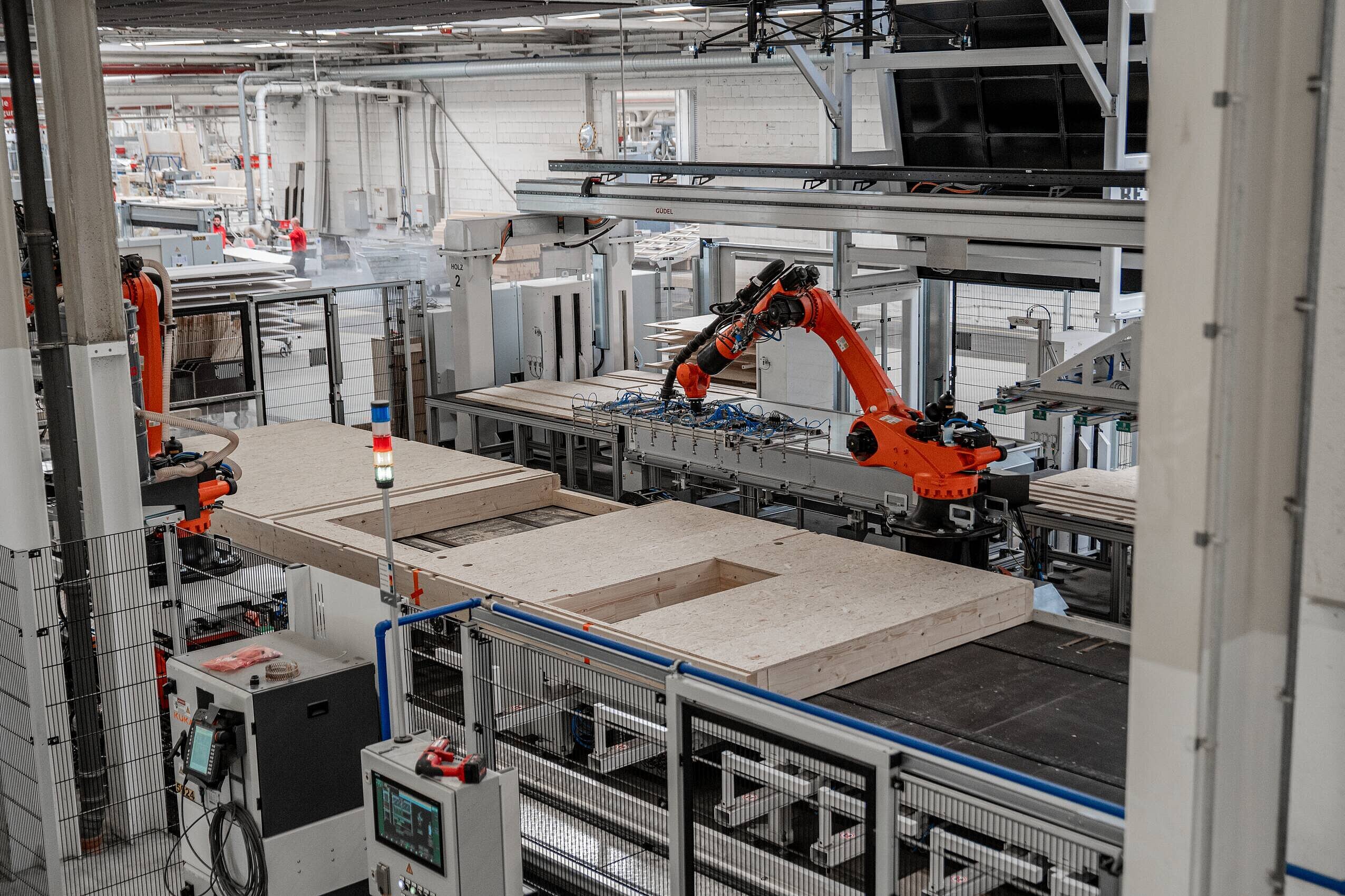 In einem modernen Produktionsraum ist ein roter Roboterarm zu sehen, der Innenwände produziert. Die Wände liegen auf einem Förderband, das für die Montage oder Bearbeitung vorgesehen ist. Im Hintergrund sind weitere Maschinen und Werkzeuge sichtbar.