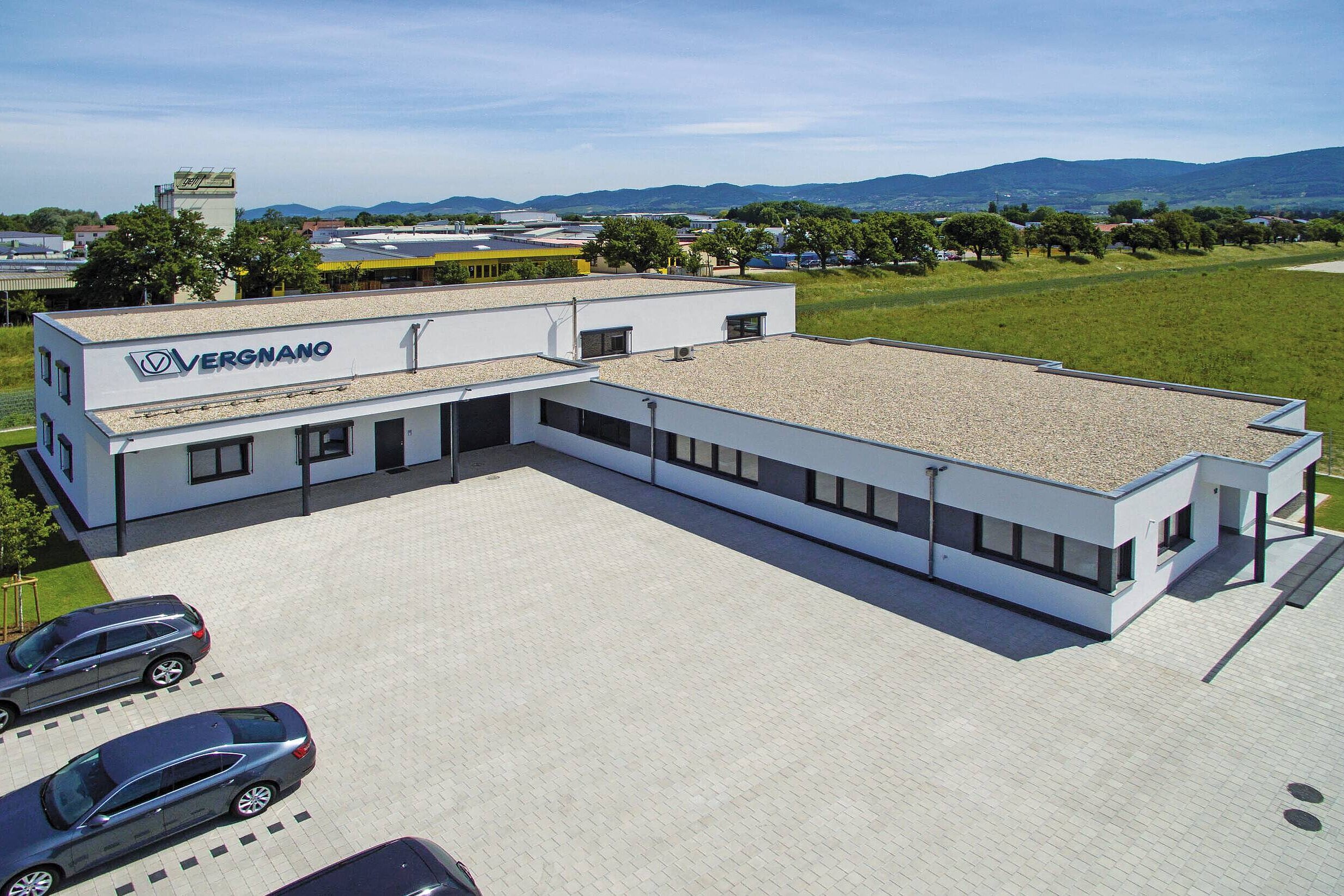 Das Bild zeigt ein Büro und Produktionsgebäude mit einem flachen Dach. Vor dem Gebäude befindet sich ein gepflasterter Parkplatz mit mehreren Autos, umgeben von einer grünen Landschaft und Hügeln im Hintergrund.