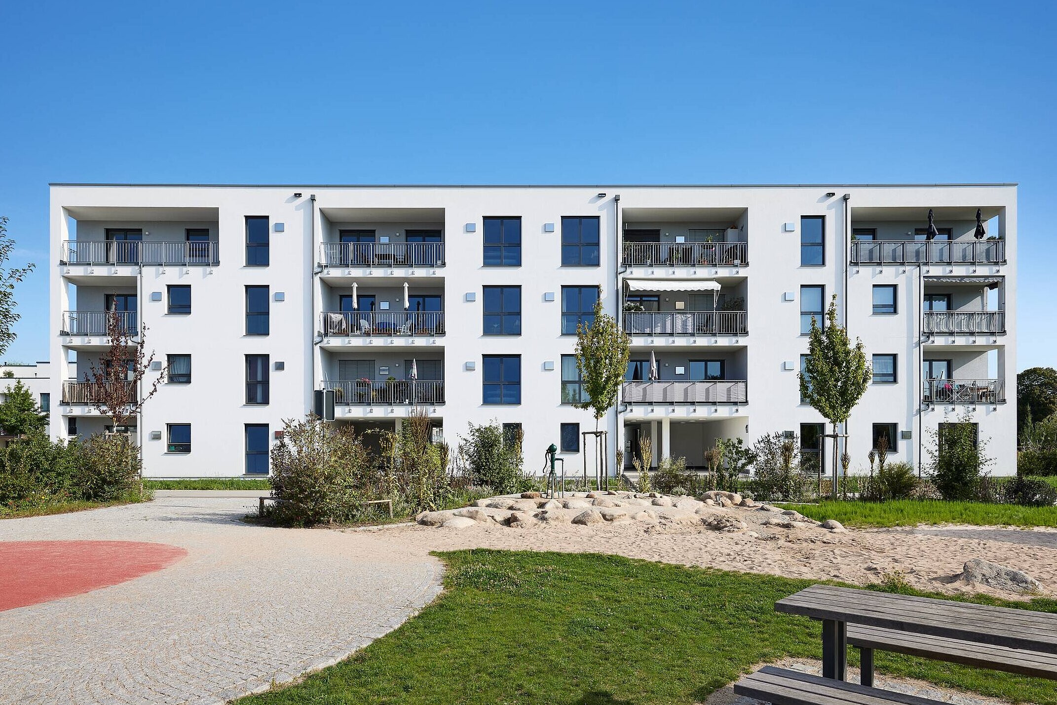 Moderner Wohnbau mit drei Etagen, großen Fenstern und Balkonen. Umgeben von grünen Flächen, Bäumen und einem Spielplatz mit Sandbereich. Der Himmel ist klar und blau.