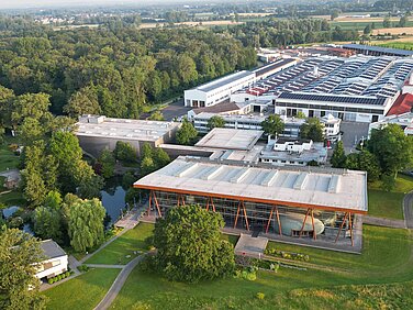 Luftaufnahme des WeberHaus Geländes, das von üppigem Grün umgeben ist. Im Vordergrund befindet sich ein modernes Gebäude mit einer großen Glasfront und Holzdetails. Im Hintergrund sind mehrere Lagerhäuser und Produktionsstätten sichtbar.