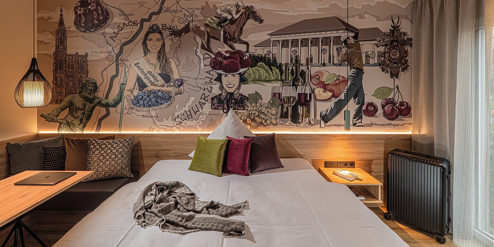 Das Bild zeigt ein stilvolles Hotelzimmer mit einer bunten Tapete, die Szenen aus der Region darstellt. Es enthält Abbildungen einer Weinlese, landwirtschaftliche Produkte, die Silhouette eines Mannes mit einer Flasche sowie eine Frau mit Trauben, die das Thema der Region feiert.
