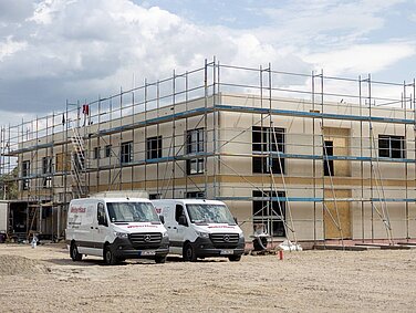 Auf dem Bild ist ein Rohbau des DRK-Gebäudes zu sehen, das mit Gerüsten umgeben ist. Im Vordergrund stehen zwei Transporter von WeberHaus, die für das Bauprojekt genutzt werden. Der Himmel ist bewölkt, und die Umgebung ist eine Baustelle mit unbefestigtem Boden.