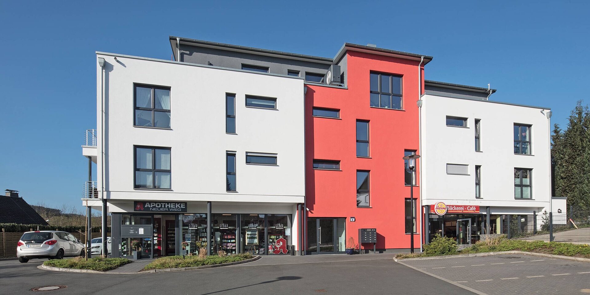 Ein modernes, vierstöckiges Gebäude mit einer weißen und roten Fassade. Im Erdgeschoss befinden sich Geschäfte, darunter eine Apotheke und eine Bäckerei. Der Himmel ist klar und blau, was den freundlichen und einladenden Charakter der Umgebung unterstreicht.