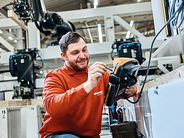 Ein Mann in einem roten Pullover arbeitet an einem technischen Gerät in einer modernen Fabrik. Er lächelt, während er mit einem Werkzeug Präzisionsarbeiten an dem Gerät durchführt. Im Hintergrund sind Maschinen und Produktionsanlagen zu sehen, die auf eine industrielle Umgebung hinweisen.