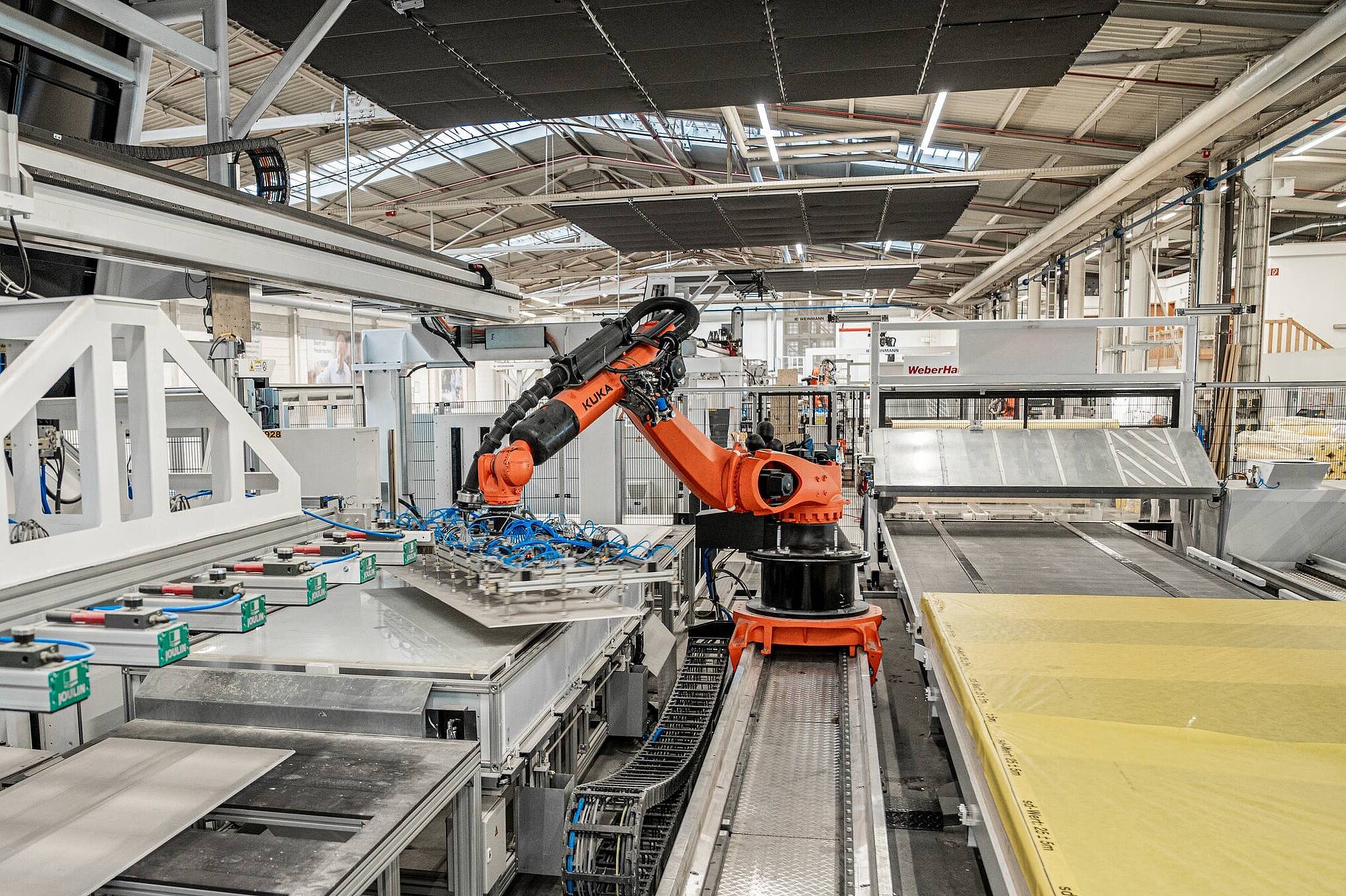 Ein industrieller Roboterarm steht in einer modernen Fertigungsanlage. Der Roboter greift eine Platte von einem Förderband, während im Hintergrund verschiedene Maschinen und Materialien sichtbar sind. Die Umgebung ist gut beleuchtet und zeigt eine effiziente Produktionsumgebung.