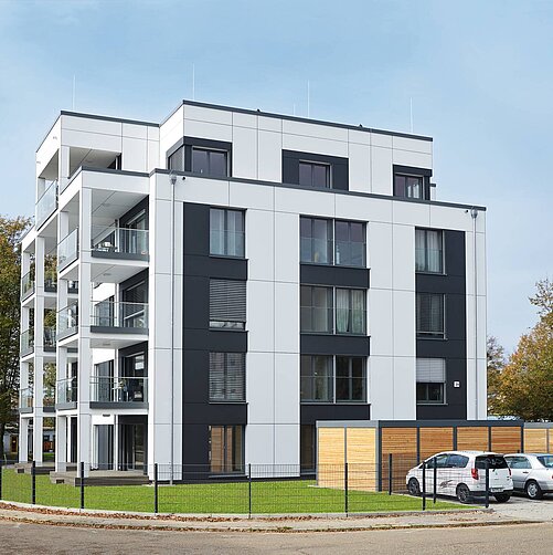 Das Bild zeigt ein modernes, Mehrfamilienhaus in einem städtischen Umfeld. Die Fassade ist in Schwarz und Weiß gehalten, mit großen Fenstern und Balkonen. Vor dem Gebäude befindet sich ein gepflegter Rasen und ein parkähnlicher Bereich mit Bäumen sowie Parkplätze.